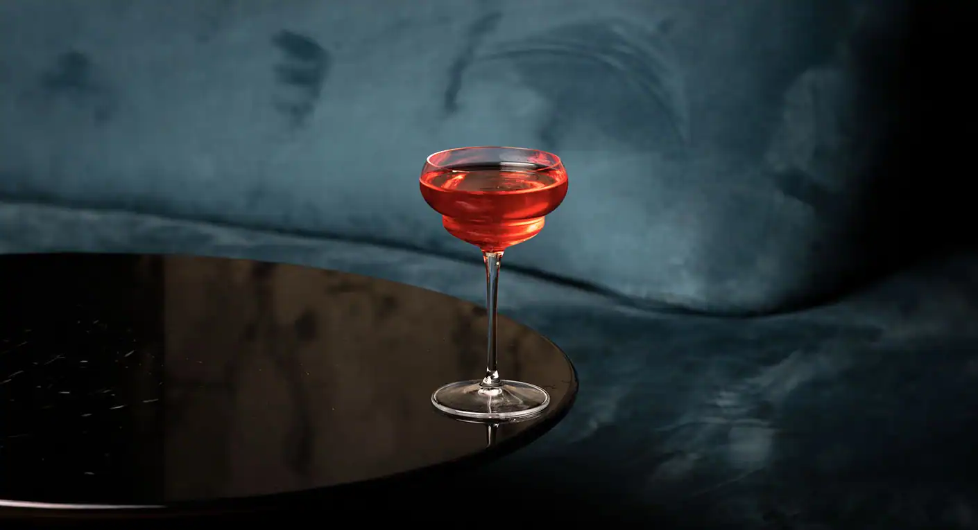 Metropolita - Signature Cocktail Cocktail rosso in calice elegante su tavolino nero riflettente, sfondo in velluto blu petrolio - Mixology Metropolita.