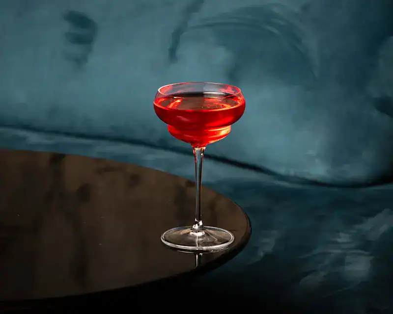 Metropolita - Signature Cocktail Cocktail rosso in calice elegante su tavolino nero riflettente, sfondo in velluto blu petrolio - Mixology Metropolita.